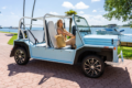 Woman driving a light blue open-air Moke vehicle along a sunny coastal waterfront promenade.